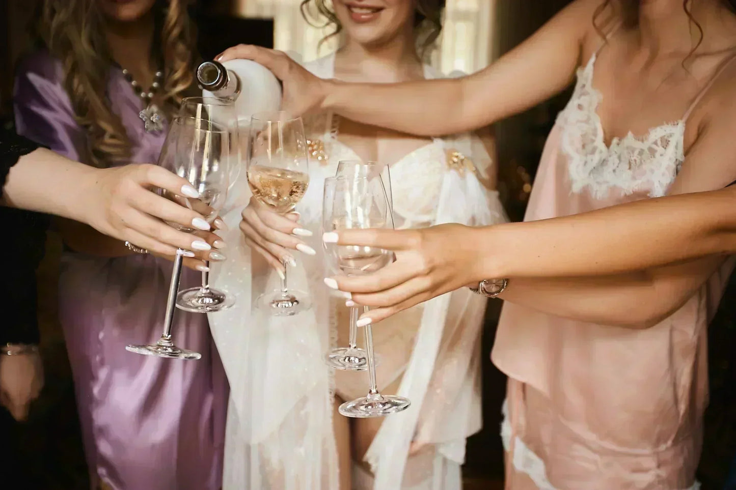 Hen party guests in satin pyjamas and robes pouring champagne into glasses during a bridal party celebration or wedding morning get-ready moment.