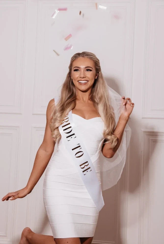 Smiling bride-to-be in a white dress and veil wearing a bridal sash with confetti falling around her.