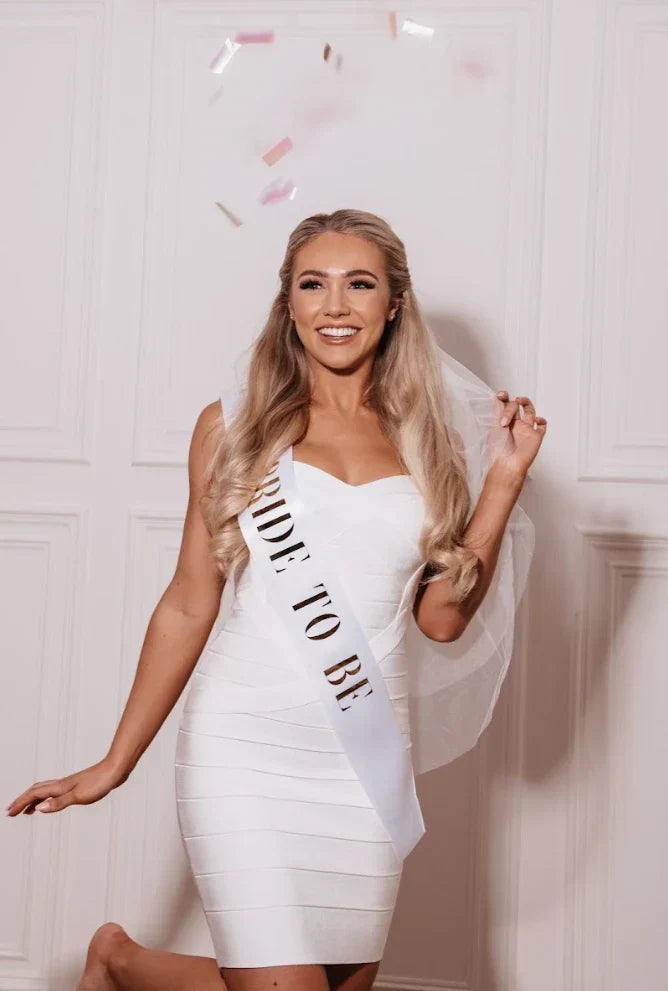 Smiling bride-to-be in a white dress and veil wearing a bridal sash with confetti falling around her.