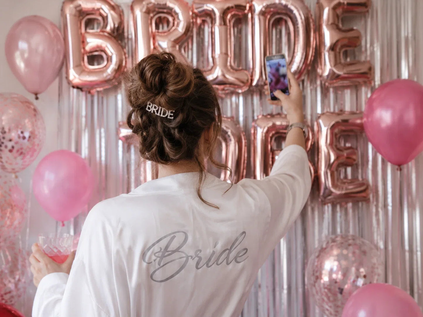 Bride in a white robe taking a photo of pink and rose gold hen party decorations before the celebration