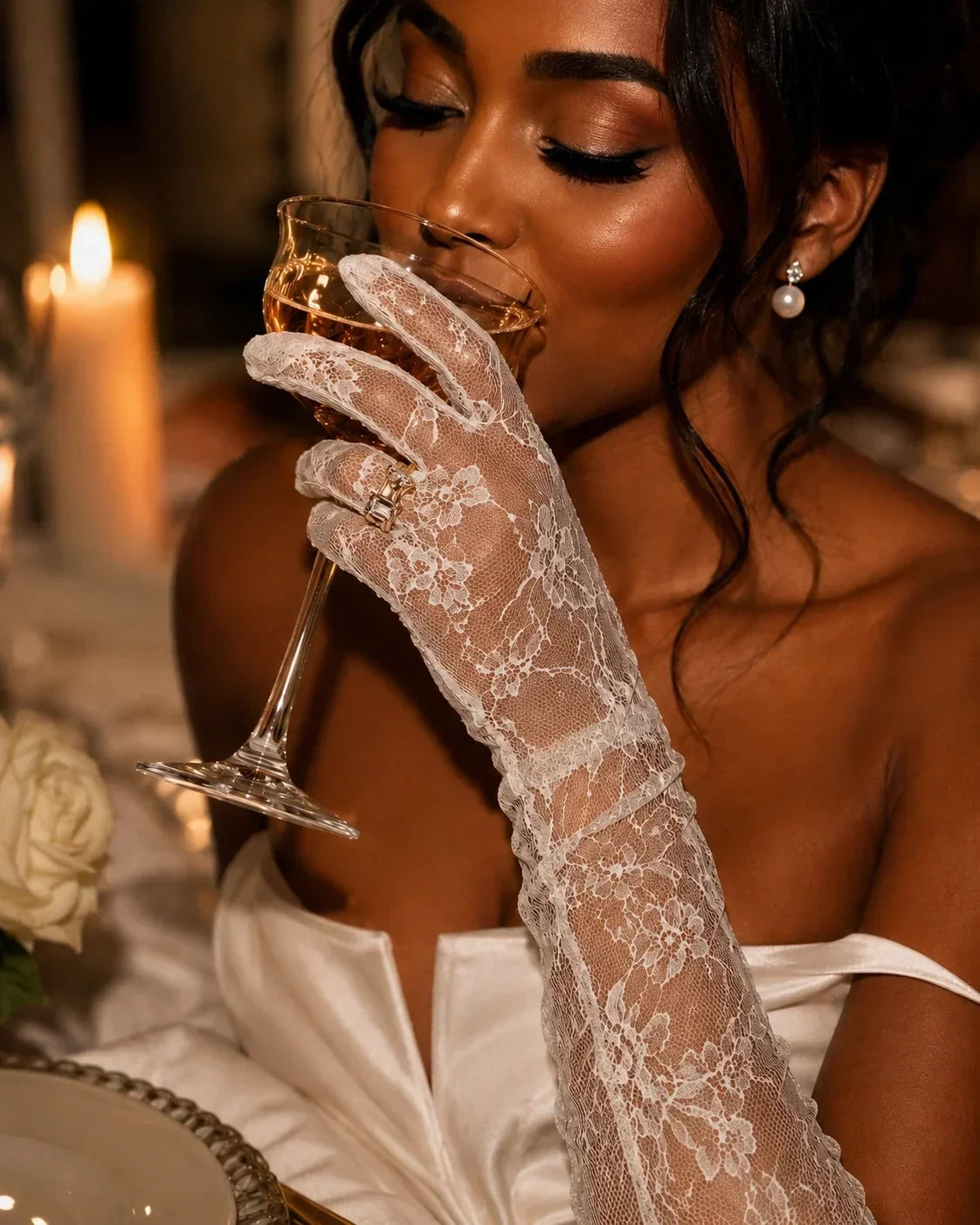 Black bride in an ivory satin gown wearing sheer white lace opera gloves, sipping champagne from a coupe glass at a candlelit reception. Pearl earrings, warm caramel-brown skin, and soft golden lighting create an elegant, luxurious bridal portrait.