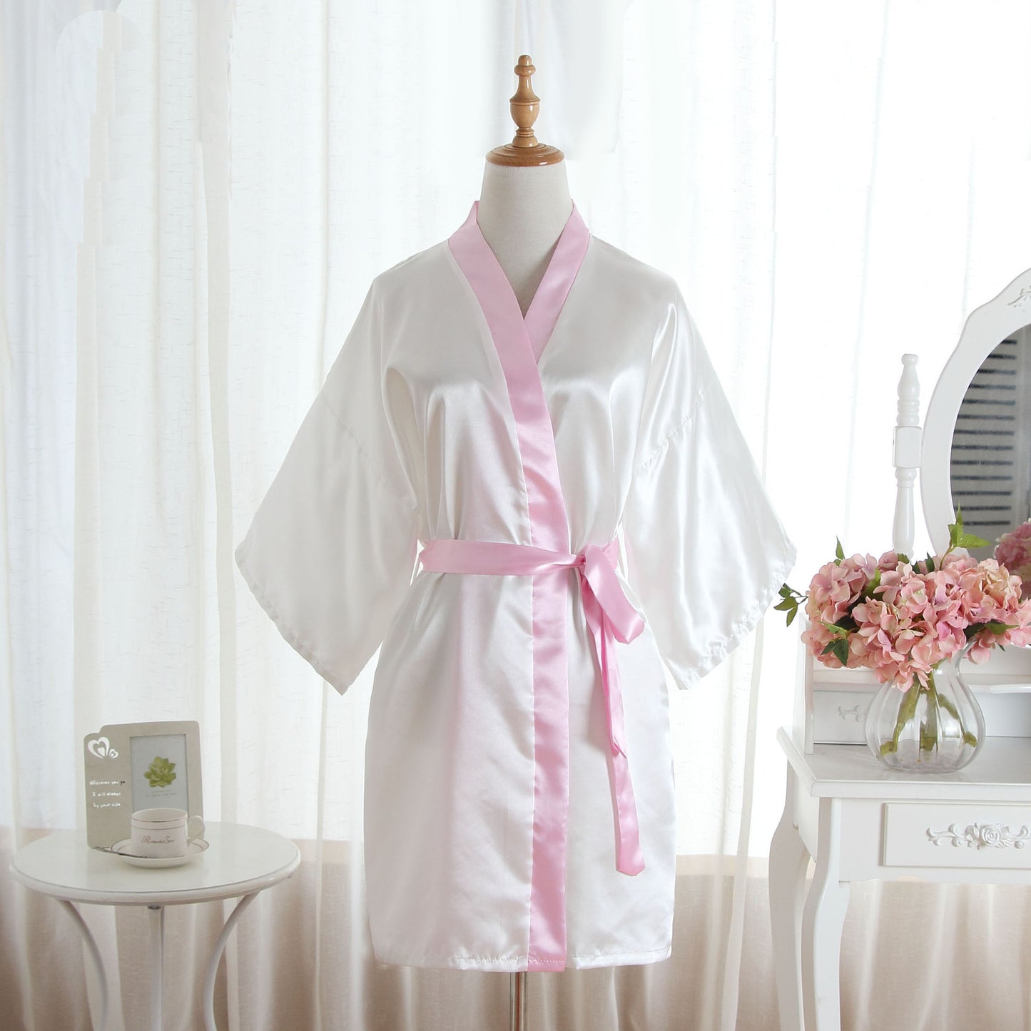 A white satin-like robe with a light pink collar, belt, and trim, displayed on a mannequin in a softly lit room.