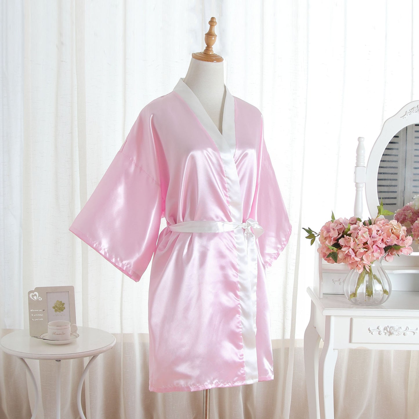 A pink satin robe with white trim and a matching white belt, displayed on a mannequin in a softly lit room with white curtains, a small table with flowers, and a mirror in the background.
