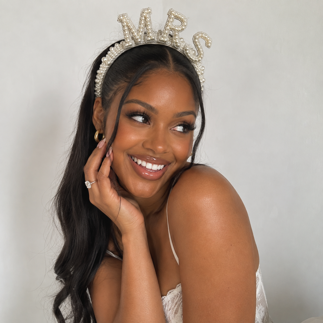 A smiling woman wearing a pearl-embellished tiara with the letters 'M-R-S' on a light gray background.