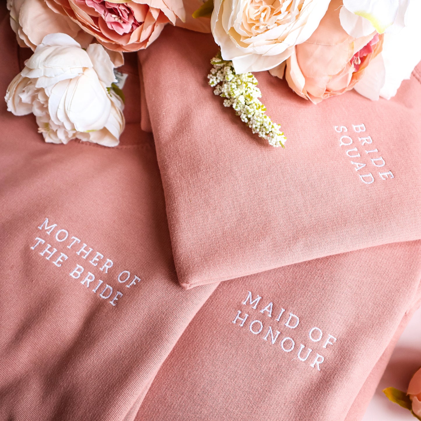 Detailed close-up of three folded dusty rose or terracotta-colored sweatshirts arranged together, each featuring white embroidered text. The top sweatshirt says "BRIDE SQUAD," the middle one says "MOTHER OF THE BRIDE," and the bottom one says "MAID OF HONOUR." A bouquet of soft cream and peach-colored peonies and white flowers rests at the top of the stack against a dark, dotted background.