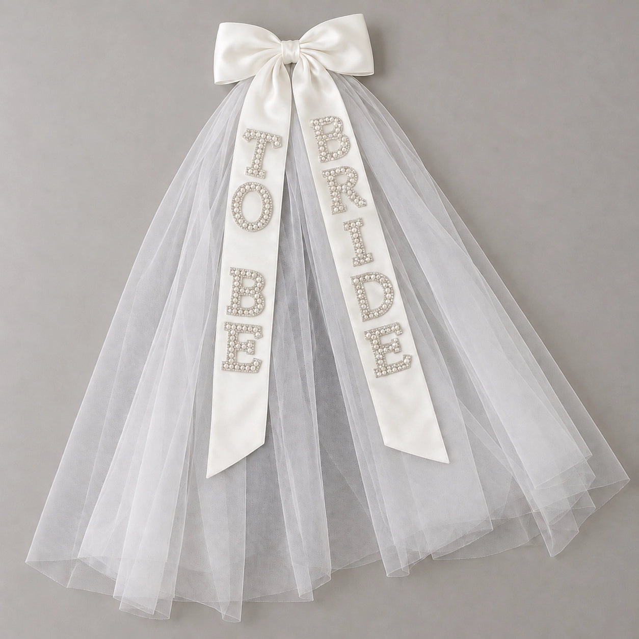 White satin bow veil with sheer tulle and pearl-effect “Bride to Be” lettering across two ribbons, displayed on a light grey background.