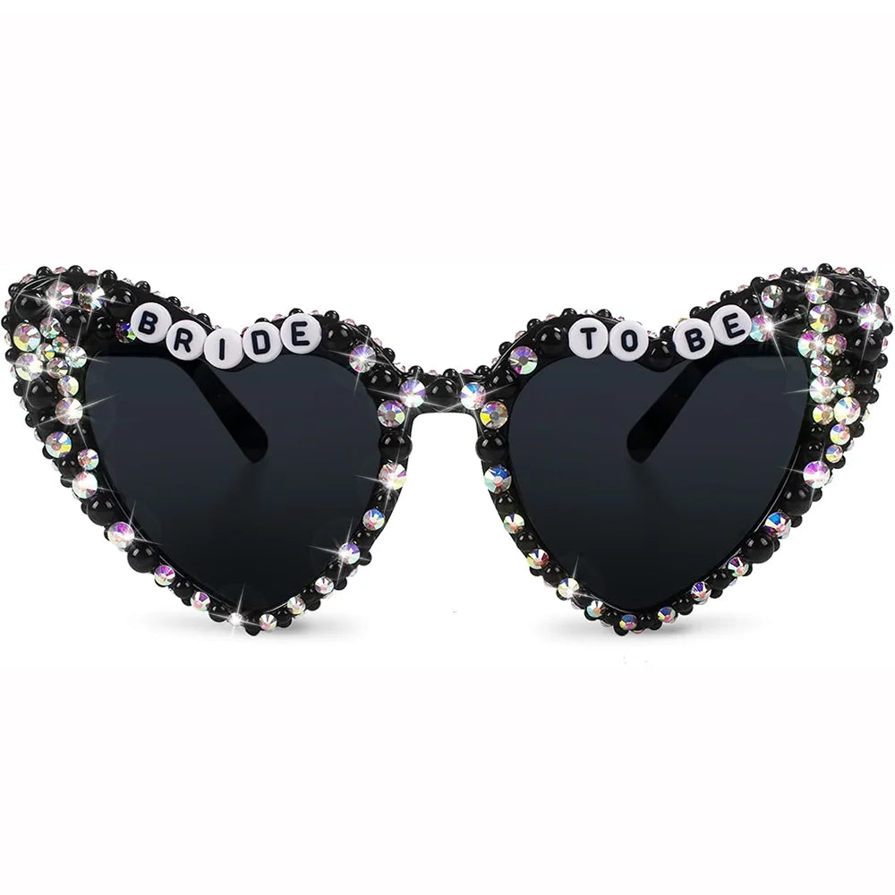 Heart-shaped sunglasses with black lenses, heavily embellished with rhinestones, and the words 'BRIDE TO BE' spelled out in white letter tiles across the bridge, shown on a plain white background.