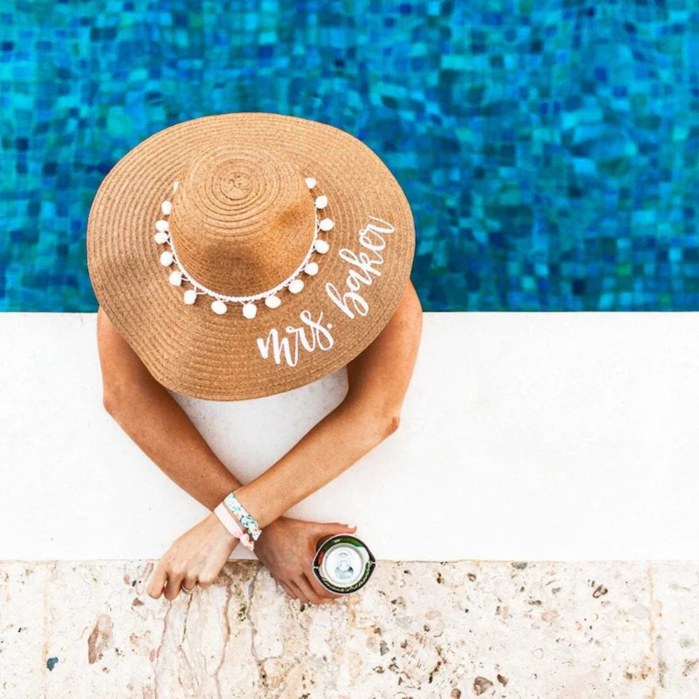 Person wearing a personalised straw hat with 'Mrs. Baker' text by a pool - The Hen Planner