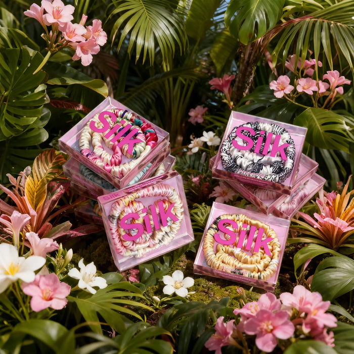 Pink boxes with 'SILK' text holding silk scrunchies surrounded by tropical flowers and leaves 