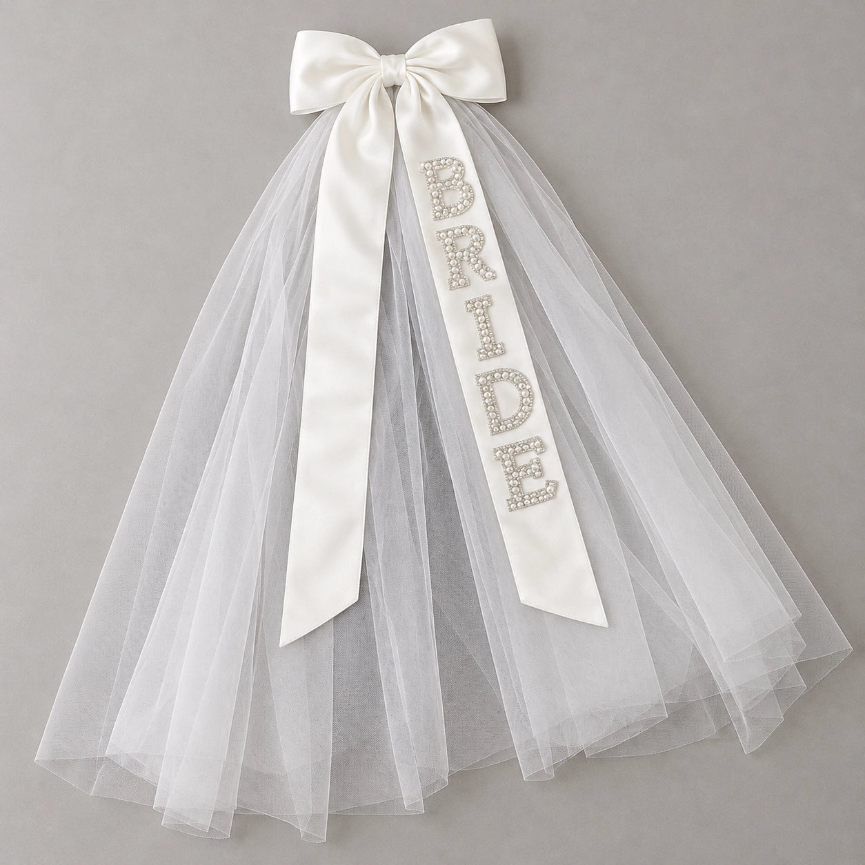 White bridal tulle veil with a large satin bow and a vertical sash spelling 'BRIDE' in rhinestone letters, shown on a neutral gray background.