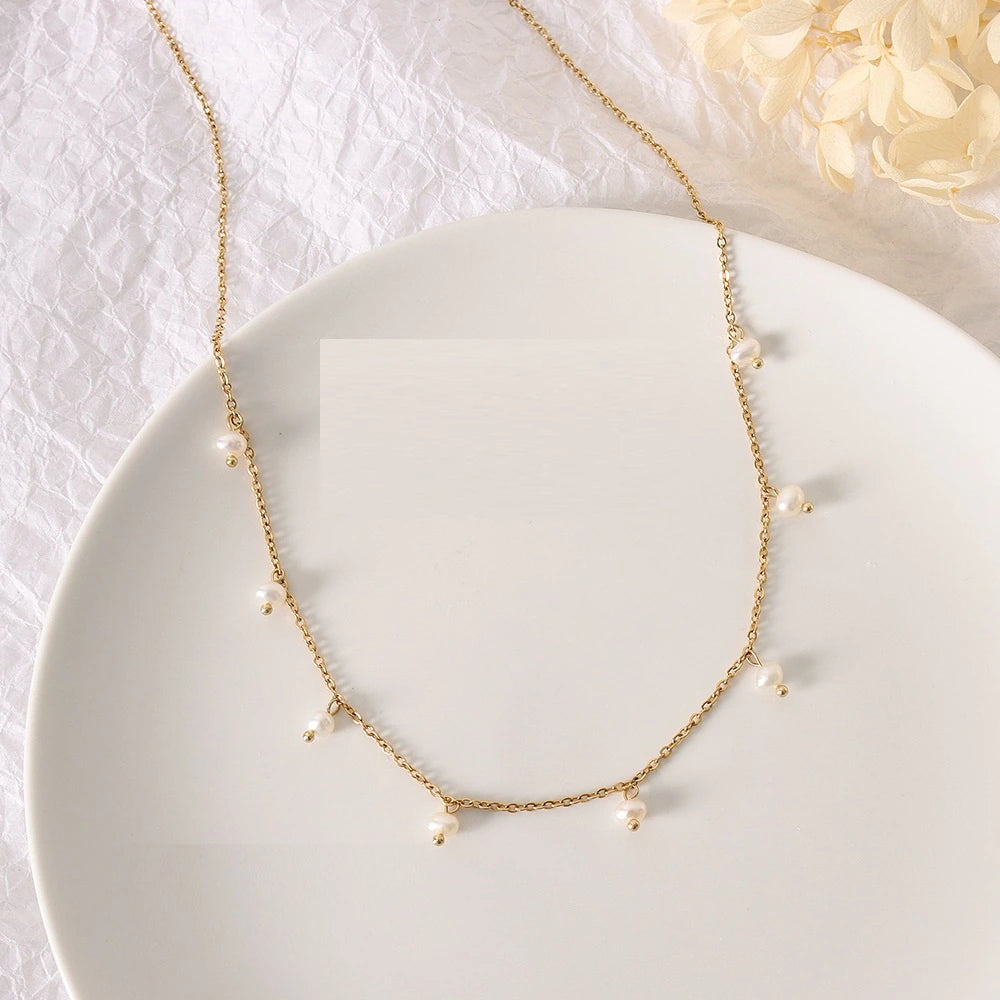 A delicate gold chain necklace with small white pearl-like beads spaced along the chain, displayed on a white oval plate against a soft white textured background with pale cream flowers in the corner.
