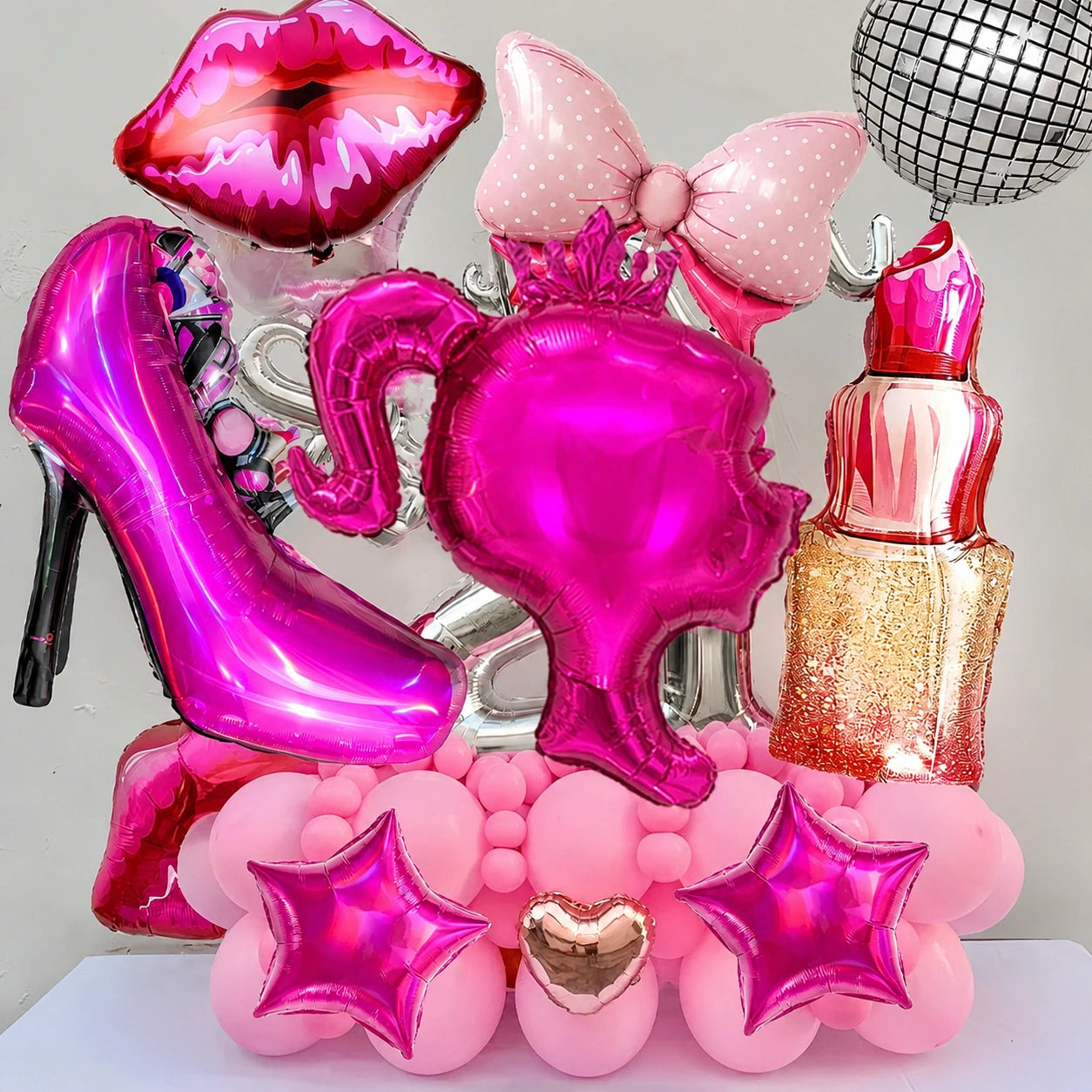 A pink and magenta birthday decoration set featuring foil balloons shaped like a high-heeled shoe, lips, bow, tiara, lipstick, star, and heart, along with pink and silver balloon balls, arranged on a white surface against a light gray background.