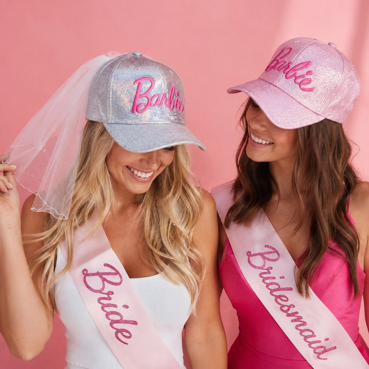 Two smiling women wearing Barbie-branded bridal accessories against a pink backdrop. One wears a silver glitter cap with a pink Barbie logo and a white veil, while the other wears a pink glitter cap.
