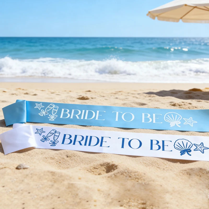 Two 'BRIDE TO BE' sashes on a sandy beach with ocean and umbrella in the background.