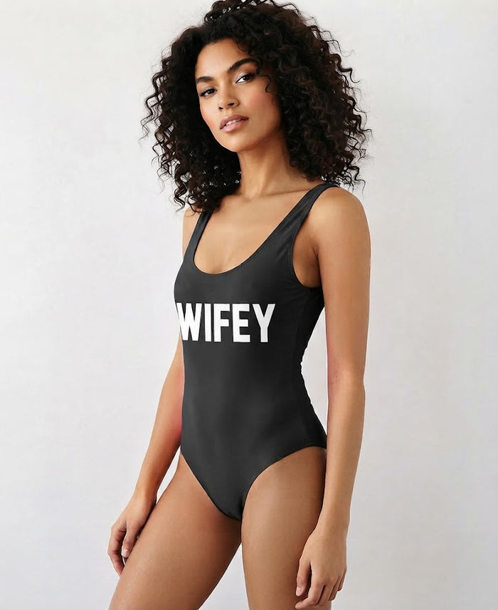 A woman modeling a black one-piece swimsuit with the word 'WIFEY' printed in bold white letters across the chest, set against a plain light gray background.