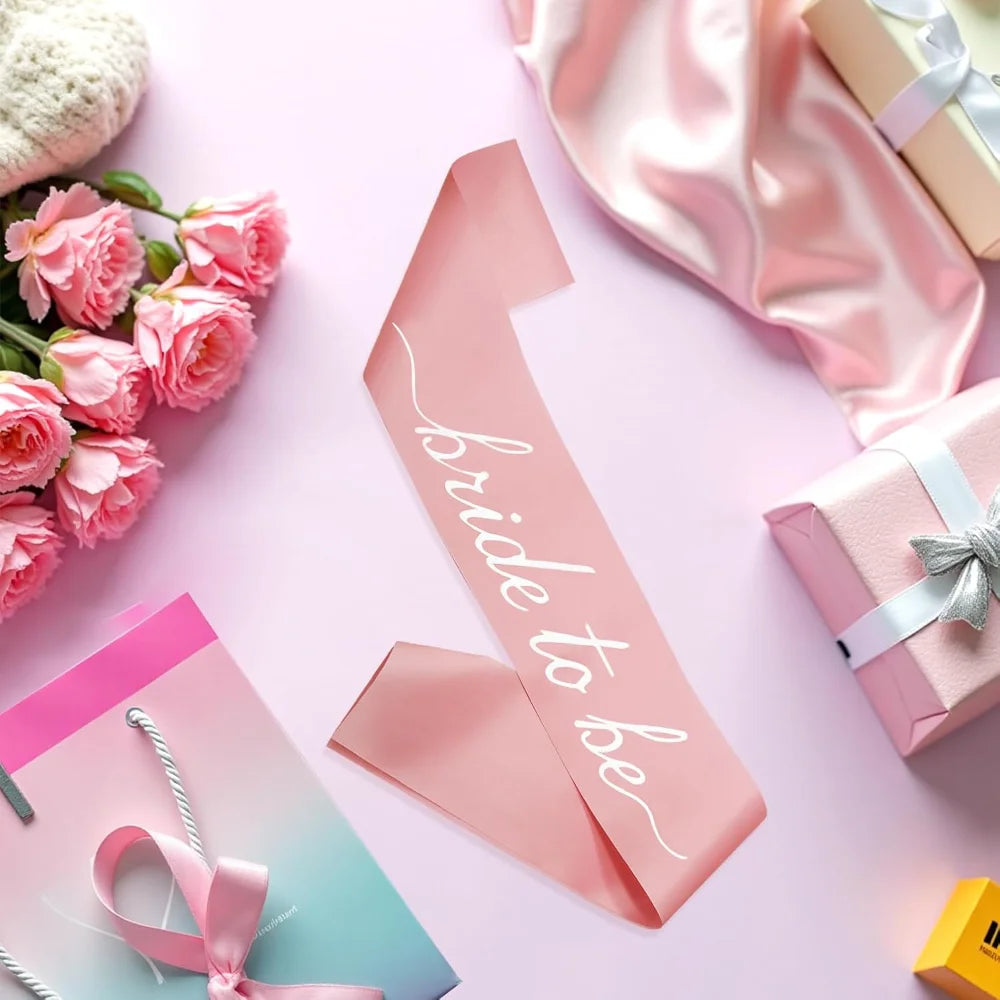 A pink satin bachelorette party sash with white script text reading 'Bride to be', styled in a flat-lay arrangement with pink roses, wrapped gift boxes, and pink satin fabric on a light pink background.