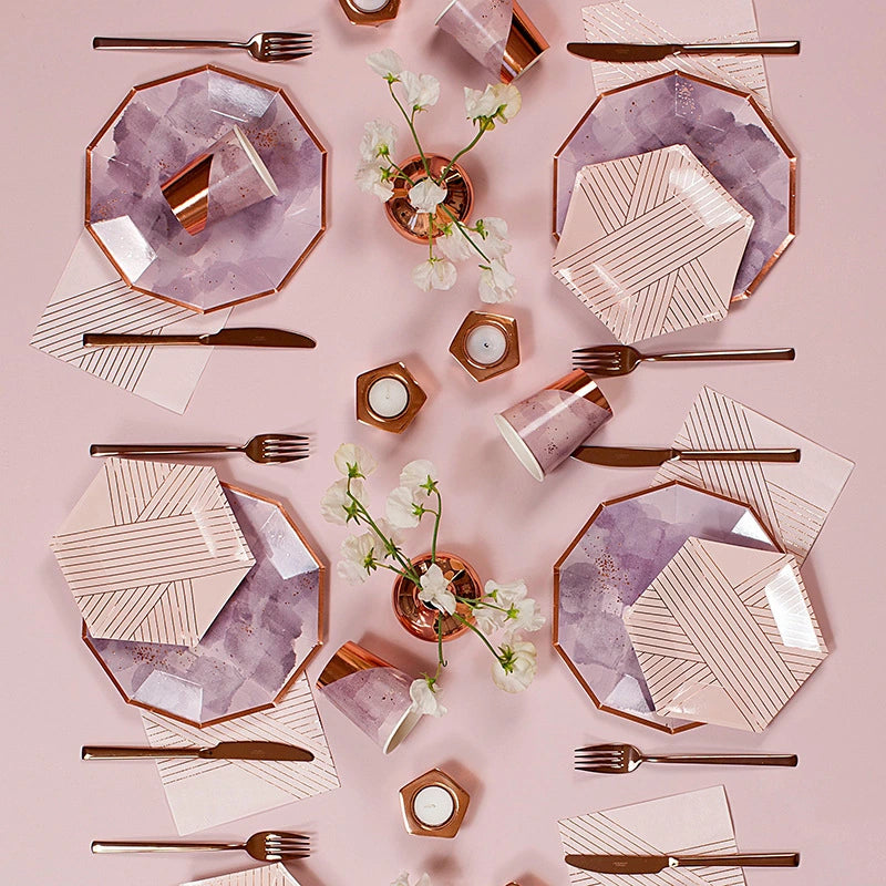 A styled flat-lay of rose gold and lavender party tableware on a dusty pink background, including octagonal plates with watercolor and striped patterns, matching paper cups, napkins, cutlery, and small floral centerpieces.