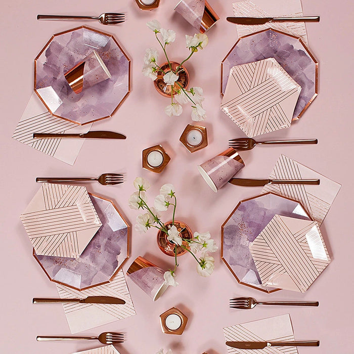 A styled flat-lay of rose gold and lavender party tableware on a dusty pink background, including octagonal plates with watercolor and striped patterns, matching paper cups, napkins, cutlery, and small floral centerpieces.