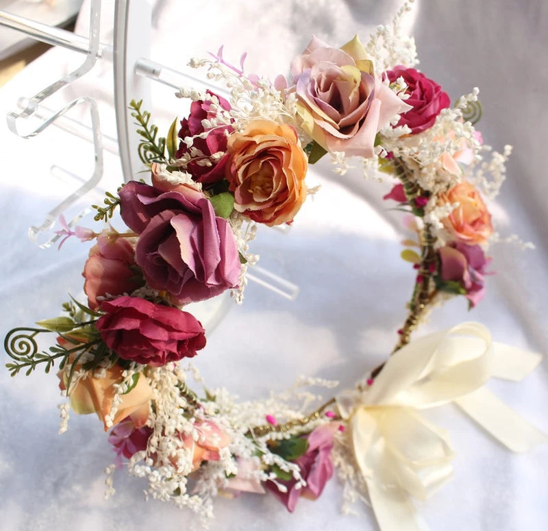 A decorative floral wreath with faux roses in pink, peach, and burgundy tones, mixed with white baby's breath and green foliage, finished with a large cream ribbon bow on a light fabric background.