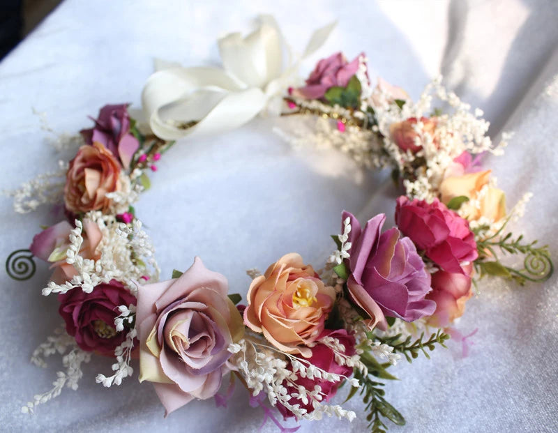 A floral crown wreath featuring dried-style roses in pink, peach, purple, and cream, accented with white baby's breath and a large white ribbon bow at the top, displayed on a white fabric background.