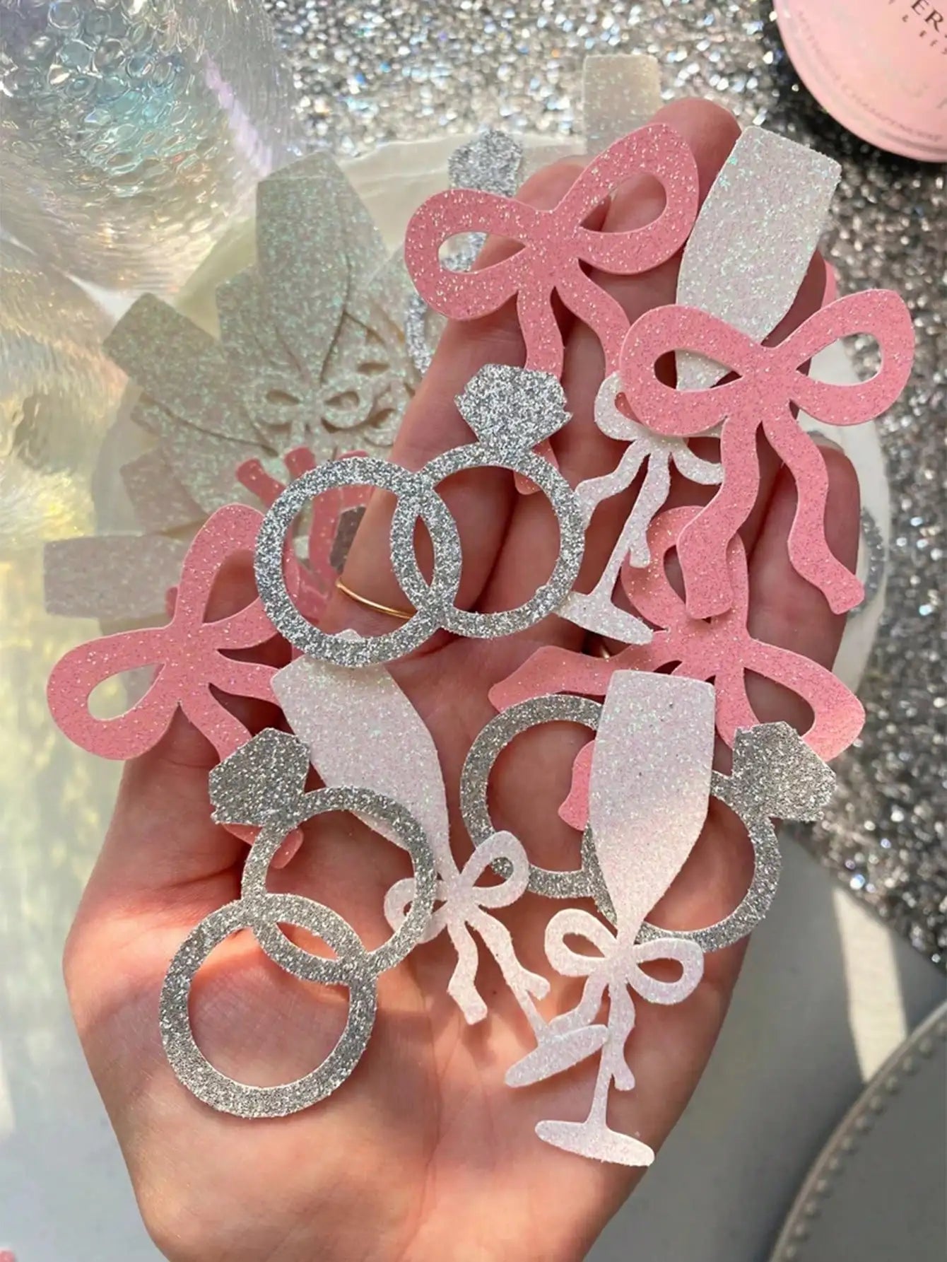 Decorative paper cutouts of rings, bows, and champagne glasses held in a hand against a glittery background.