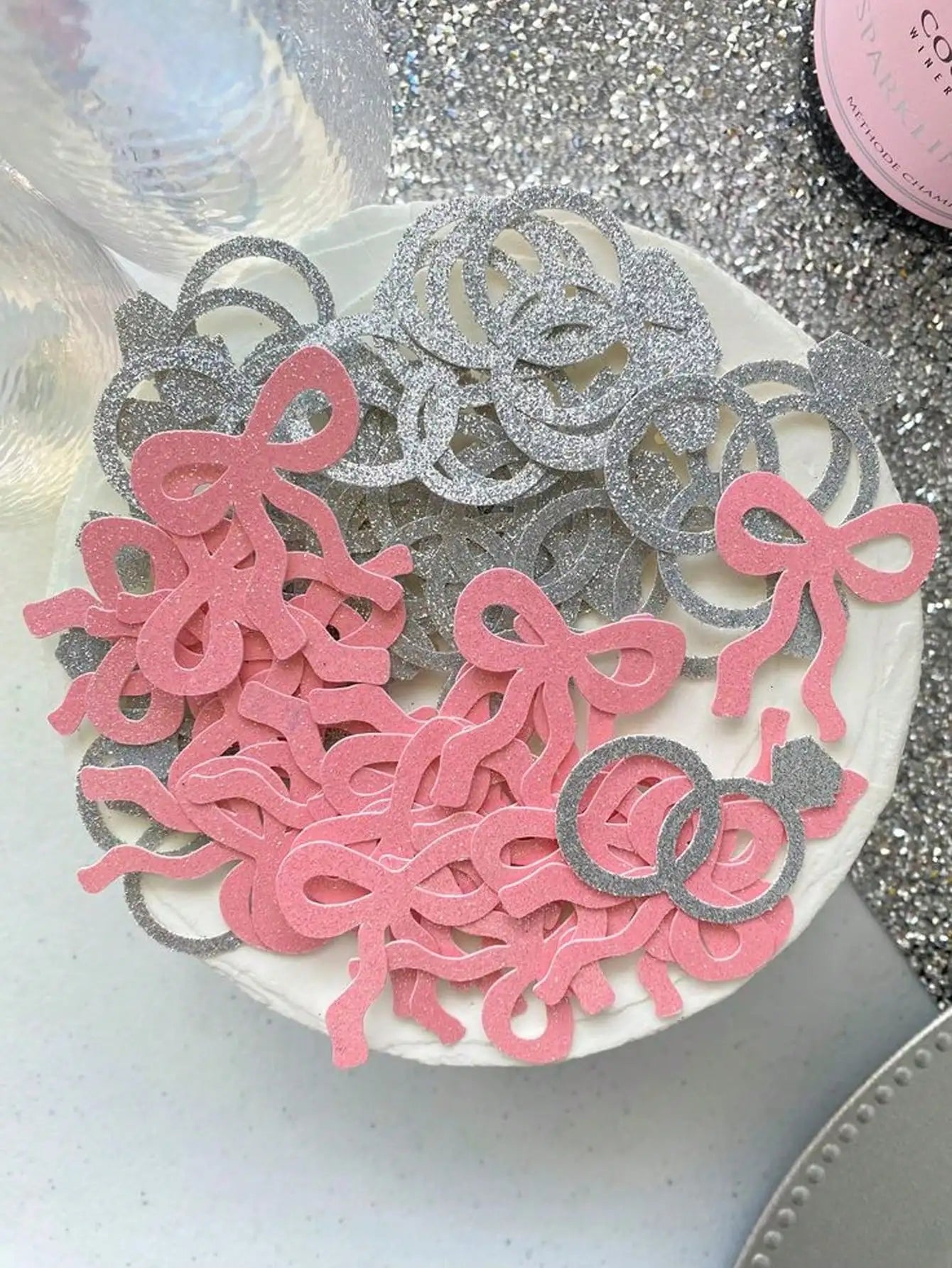 Decorative confetti with pink ribbons and silver rings on a white plate against a glittery background.