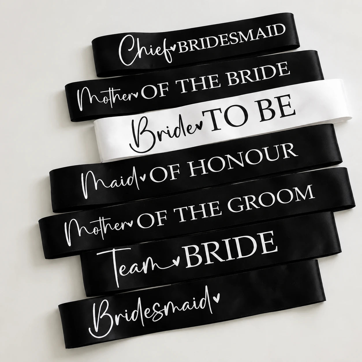 Black and white bridal party sashes laid flat on a plain white background, featuring Bride to Be, Team Bride, Bridesmaid, Maid of Honour, Mother of the Bride, Mother of the Groom, and Chief Bridesmaid designs.