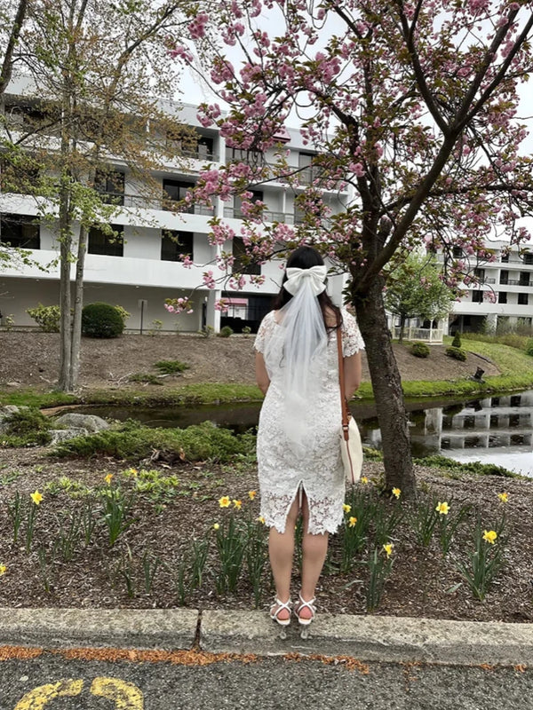 Bride to be in a white dress standing under a cherry blossom tree with a building in the background.