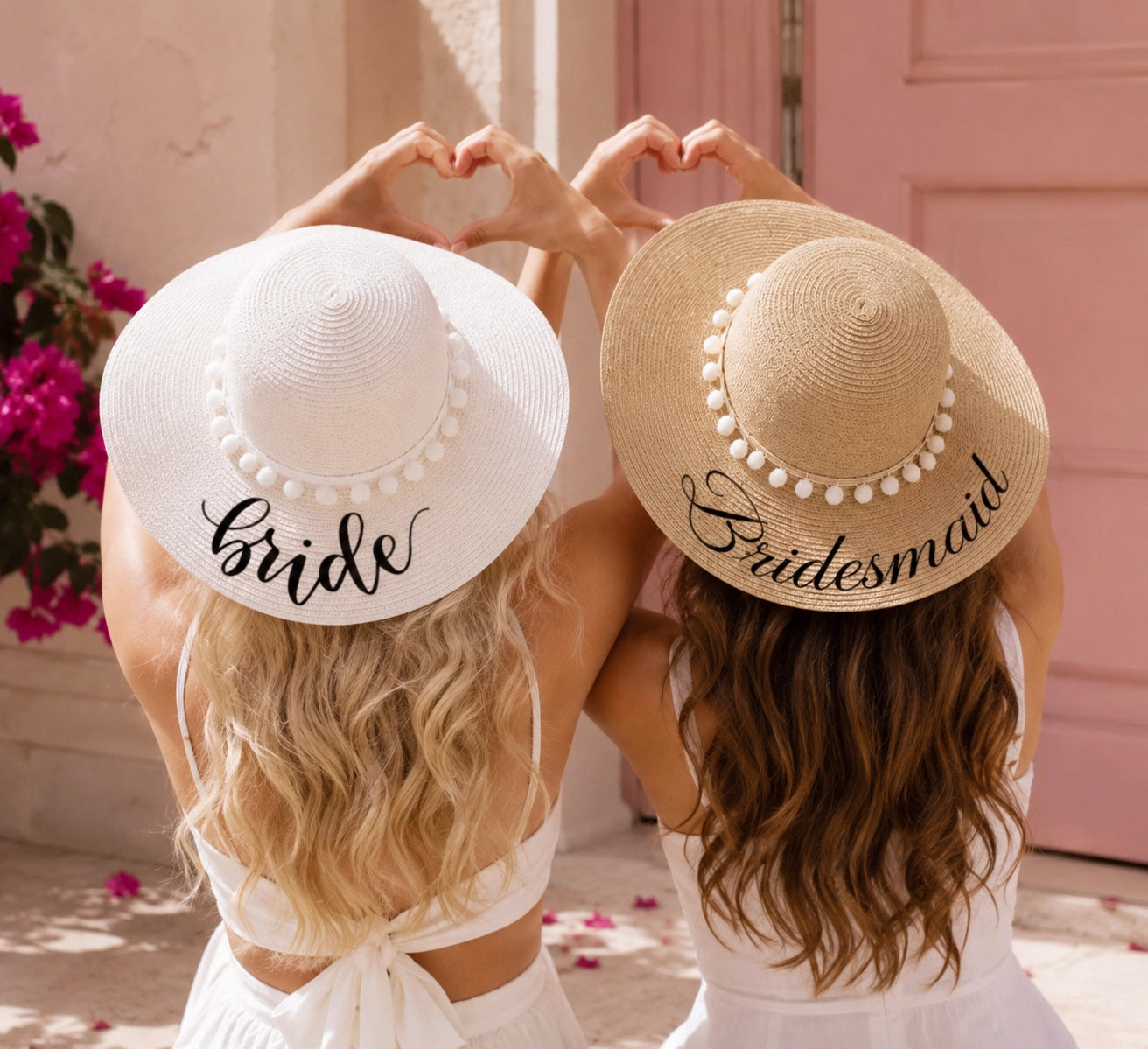 Two women seen from behind wearing wide-brimmed straw and white sun hats with decorative pearl trim and the words 'Bride' and 'Bridesmaid' in script. They are posing with their hands forming a heart shape above their heads in front of a pink door and pink flowers.