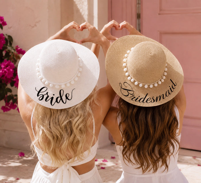 Two women seen from behind wearing wide-brimmed straw and white sun hats with decorative pearl trim and the words 'Bride' and 'Bridesmaid' in script. They are posing with their hands forming a heart shape above their heads in front of a pink door and pink flowers.