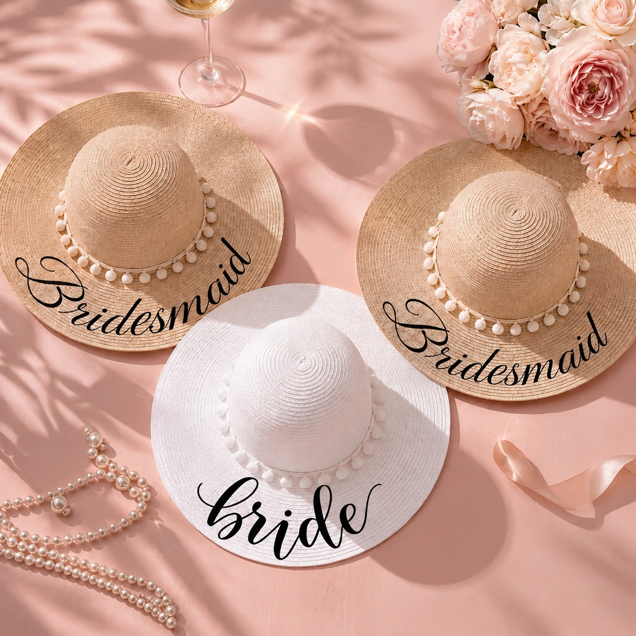 Bride and bridesmaid personalized wide-brim sun hats arranged on a soft pink backdrop with blush flowers, champagne, pearls, and ribbon, featuring two natural straw hats labeled "Bridesmaid" and one white hat labeled "Bride."
