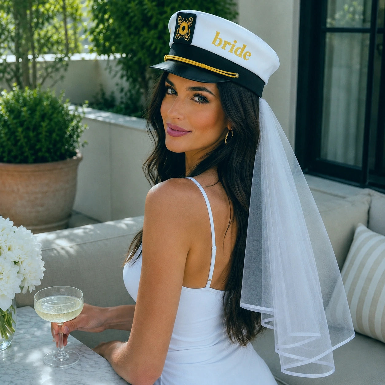 A woman wearing a white bridal sailor hat with a matching tulle veil sits on a patio. The hat is white with a black band, gold trim, and the word 'Bride' in gold lettering, featuring a small sun-and-moon emblem on the front.