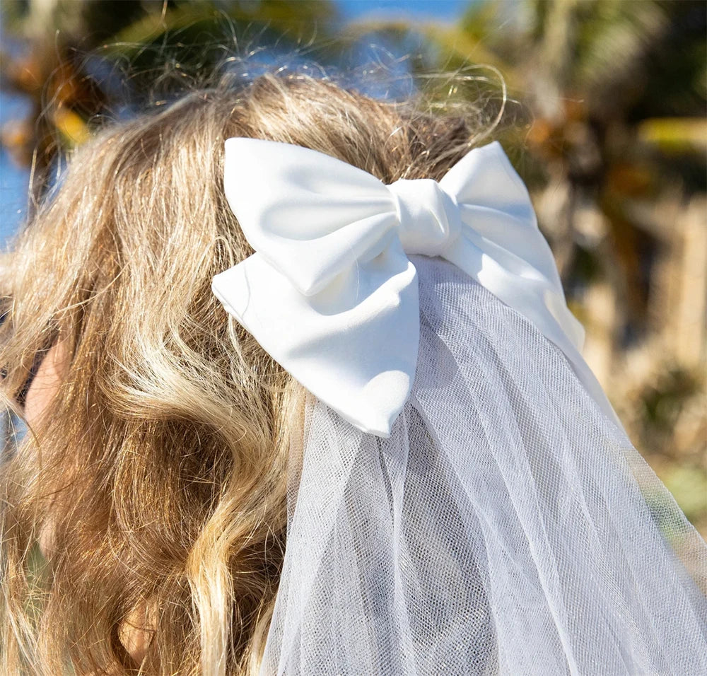 White bow with a veil attached to blonde hair against a blurred natural background