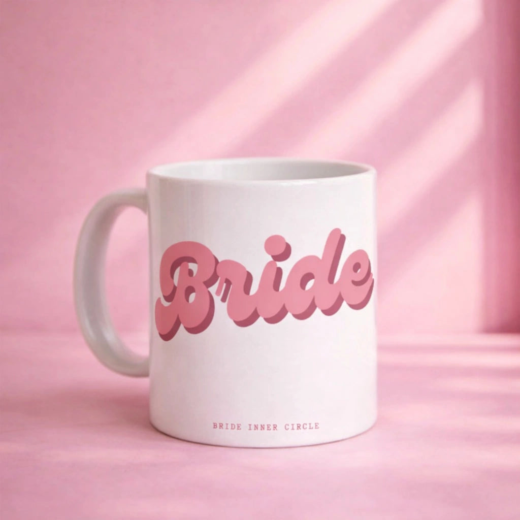 White ceramic mug with pink retro-style “Bride” lettering on the front, styled on a soft pink background with natural light and subtle shadow details.