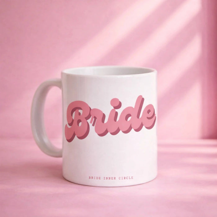 White ceramic mug with pink retro-style “Bride” lettering on the front, styled on a soft pink background with natural light and subtle shadow details.