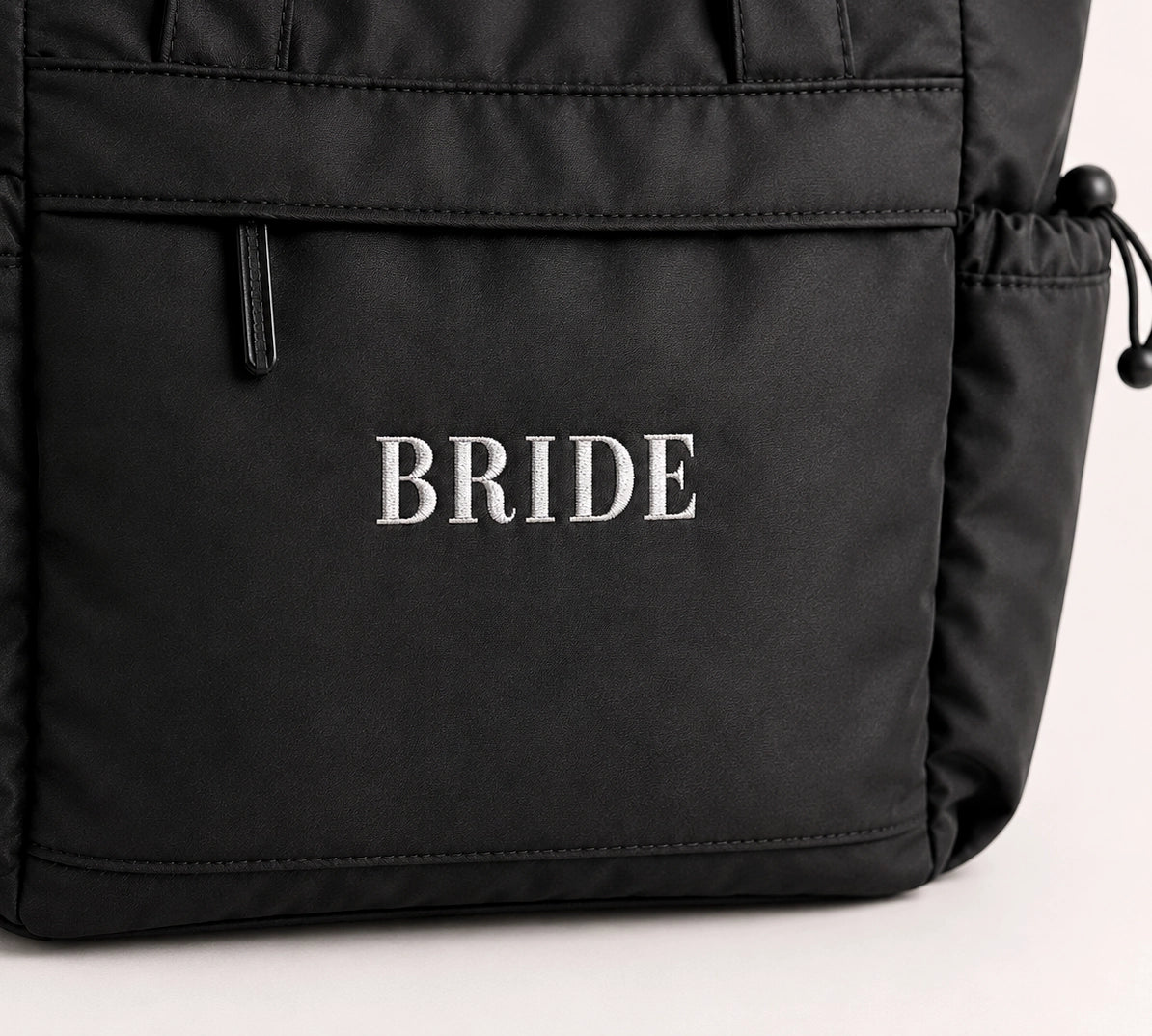 Close-up of a black nylon bag with the word 'BRIDE' embroidered in white serif letters on the front pocket.