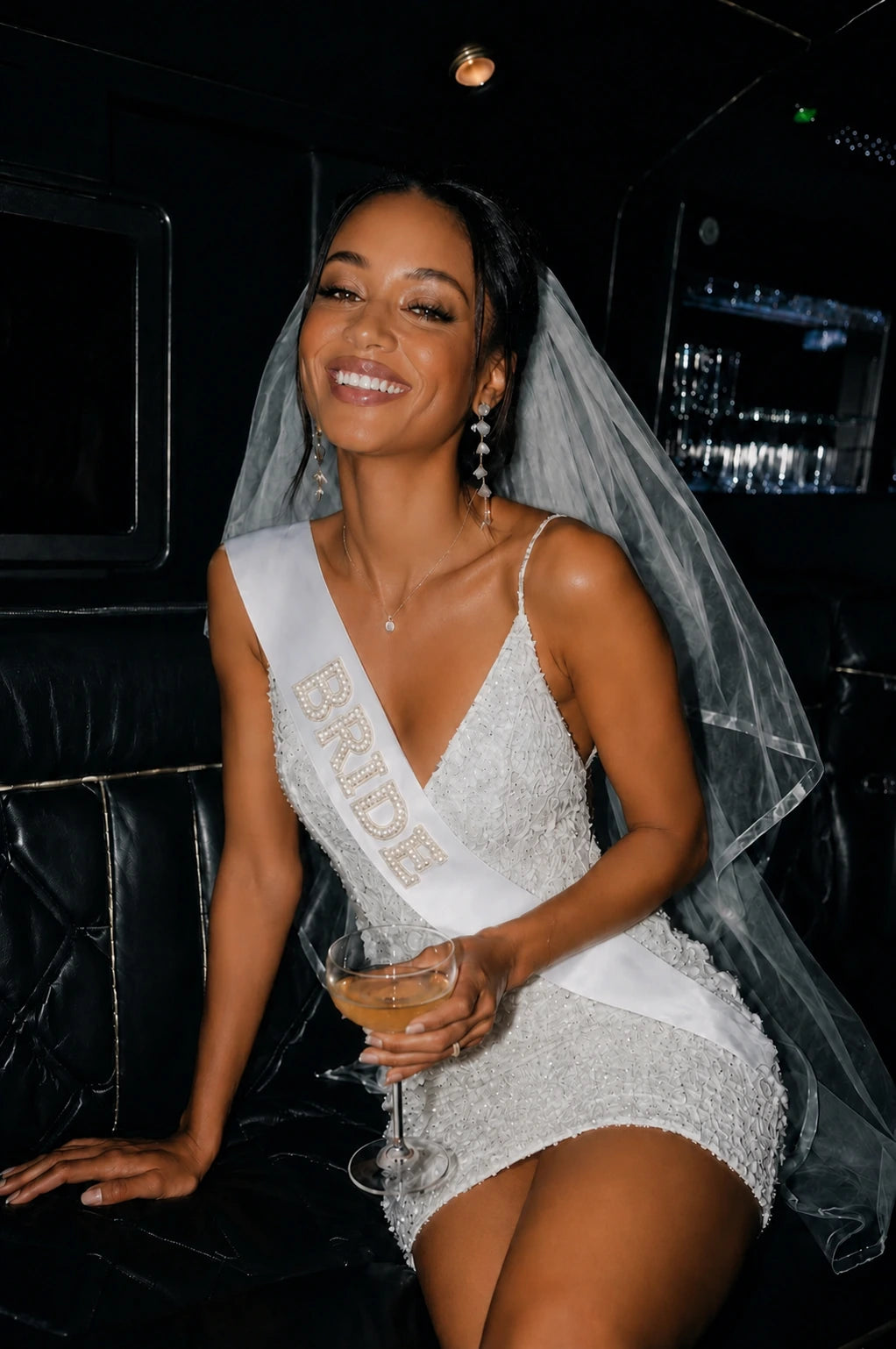 Woman in a white dress with 'Bride' sash holding a glass, in a limousine.
