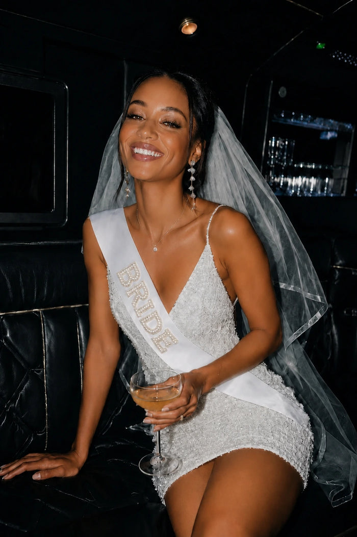 Woman in a white dress with 'Bride' sash holding a glass, in a limousine.