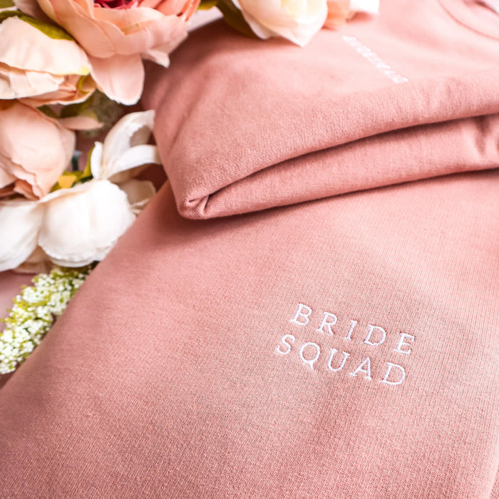 A close-up, angled shot of a folded dusty rose sweatshirt with "BRIDE SQUAD" embroidered in white serif capital letters. Another folded sweatshirt is partially visible in the background, along with soft peach and cream peonies to the left, creating a clean and elegant aesthetic.