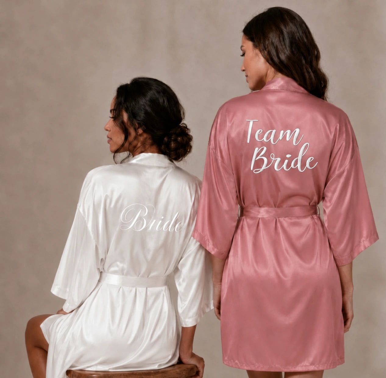 Bride to be wearing a white robe with white scribe saying 'Bride' and a female standing next to her wearing a rusty pink robe with 'Team Bride'