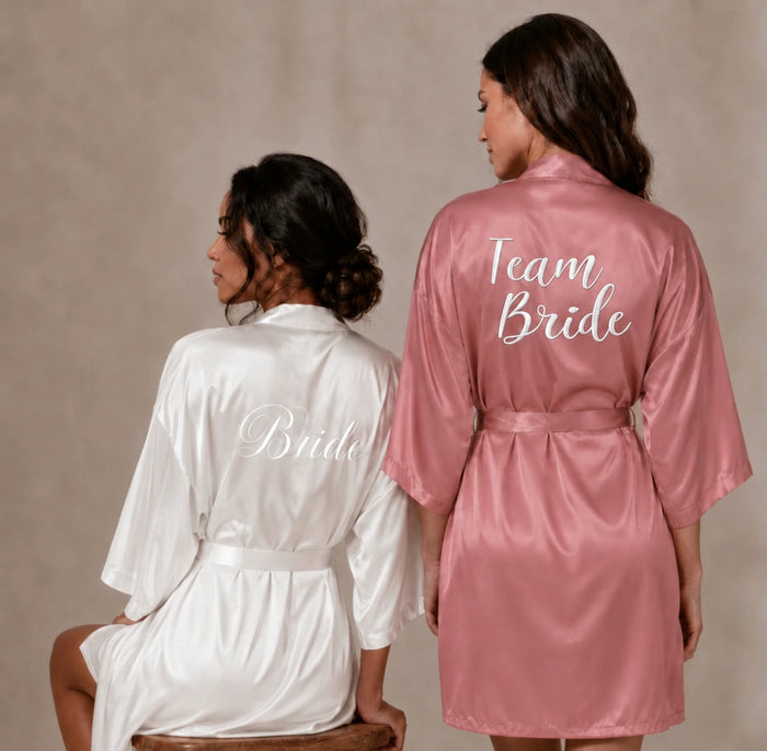 Bride to be wearing a white robe with white scribe saying 'Bride' and a female standing next to her wearing a rusty pink robe with 'Team Bride'