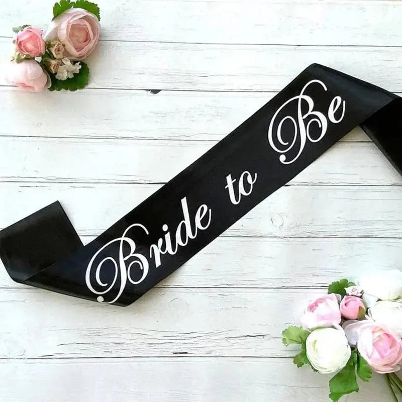 Black sash with 'Bride to Be' text on a white wooden background with flowers.
