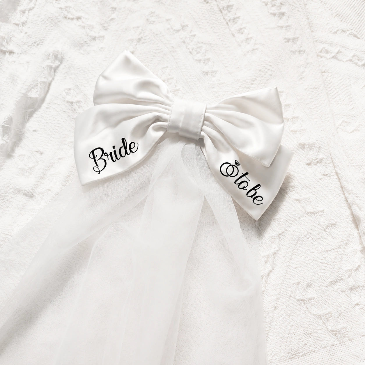 A white satin and tulle bridal hair bow with 'Bride' on one side and 'Stake' on the other, displayed on a textured white lace fabric background.