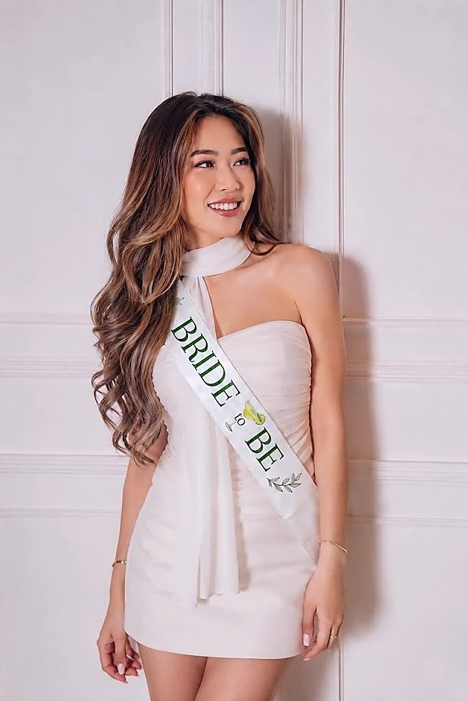 Woman wearing a white dress and 'Bride to Be' sash against a light-colored wall.
