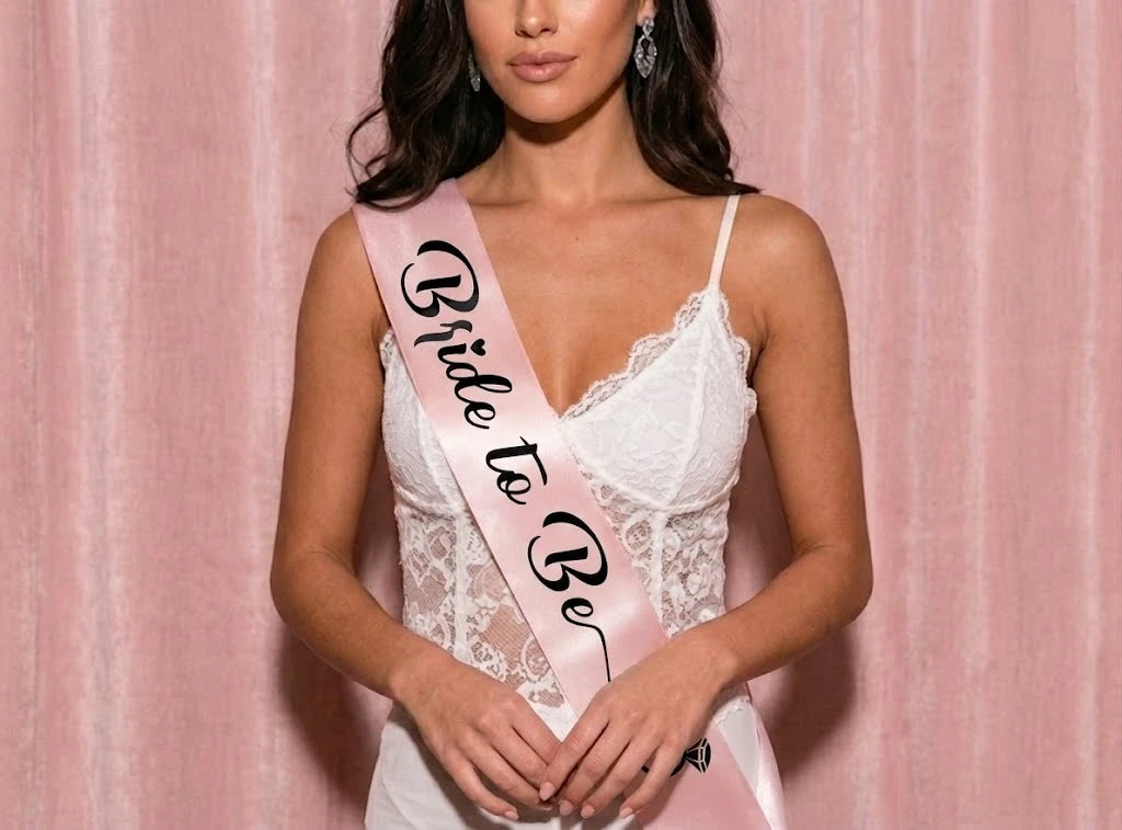 Woman wearing a white lace dress and 'Bride To Be' sash against a pink background
