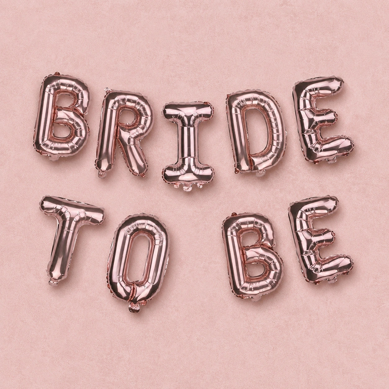 Rose gold foil balloon letters spelling out 'BRIDE TO BE' arranged in two lines on a light pink background.