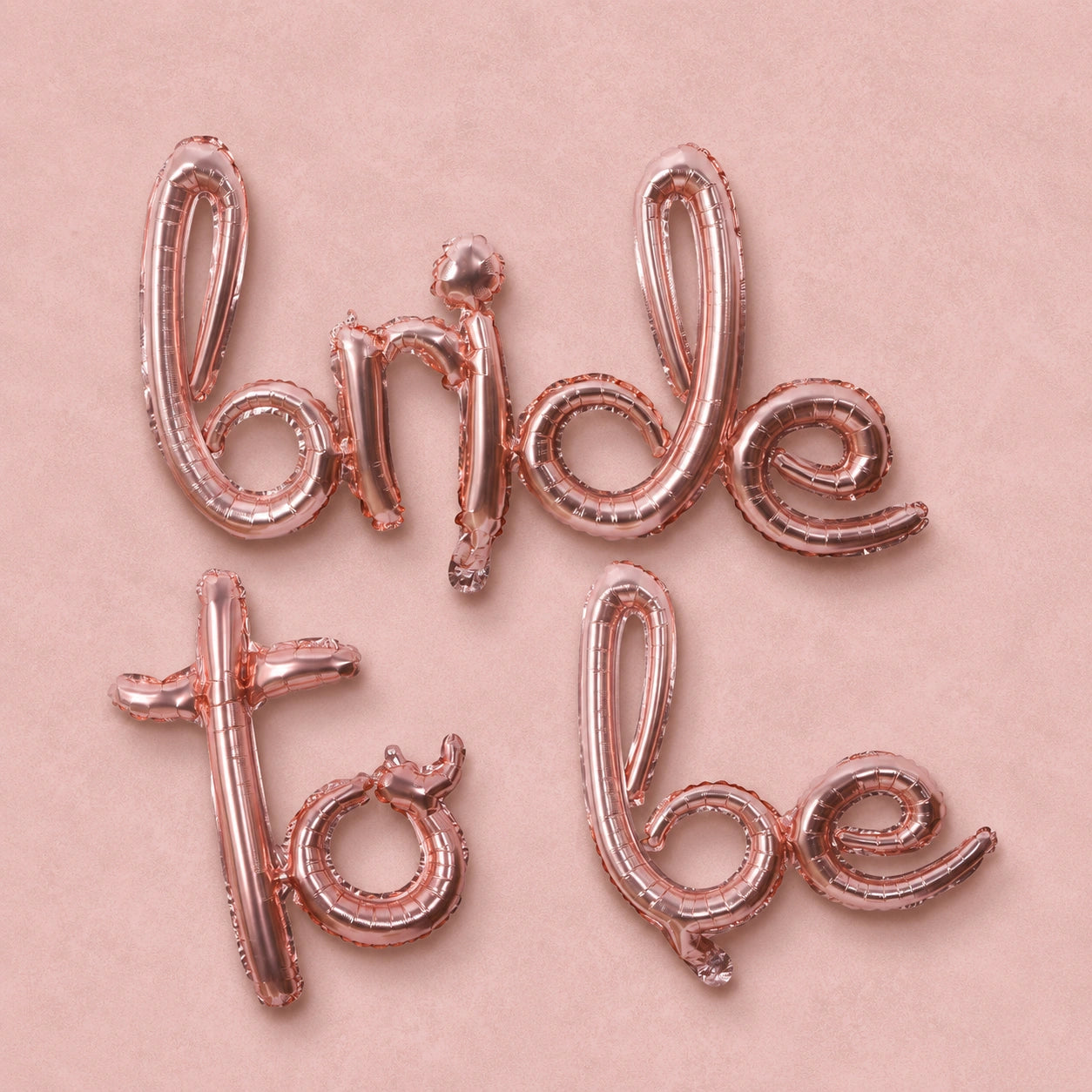 Rose gold foil balloon letters spelling 'Bride to be' arranged on a light pink background.