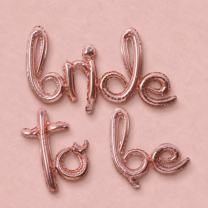 Rose gold foil balloon letters spelling 'Bride to be' arranged on a light pink background.