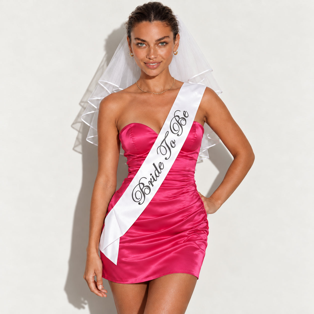 Model in a pink satin mini dress wearing a white Bride To Be sash and two-tier ribbon-edged veil.