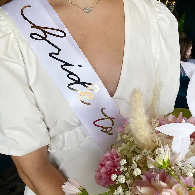 Person wearing a 'Bride To Be' sash holding flowers in an indoor setting